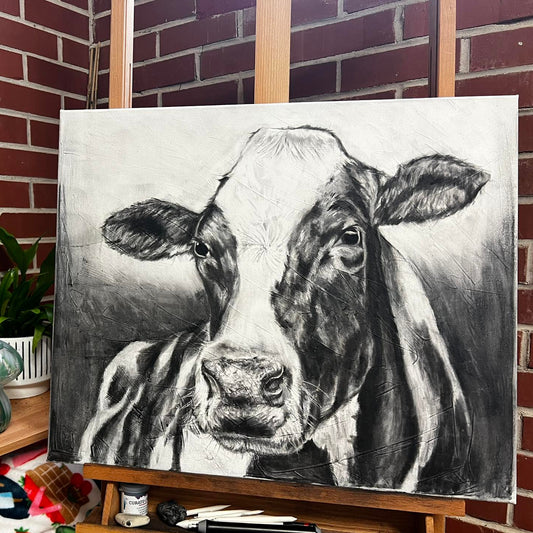 Canvas with a black and white cow painting on an easel against a brick wall.